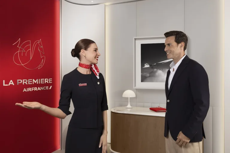 Accueil de l’espace La Première d’Air France à Paris-CDG, une agente en uniforme accueillant un voyageur devant le panneau rouge La Première / Air France La Première lounge reception at Paris-CDG, staff welcoming a traveller beside the red La Première sign.